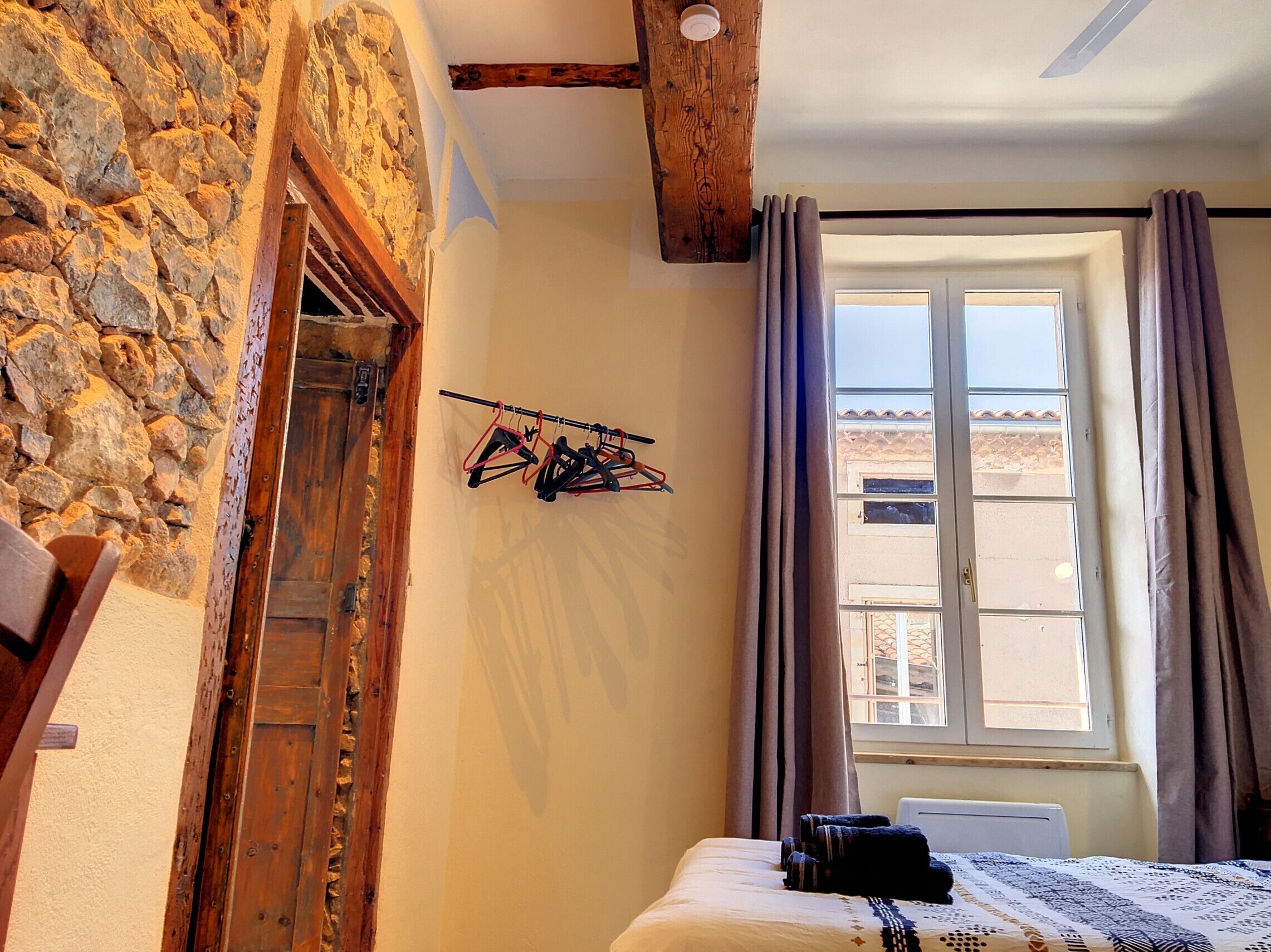 Una habitación con pared de piedra, vigas de madera, una cama con colcha estampada, una ventana con cortinas y un tendedero con ropa.