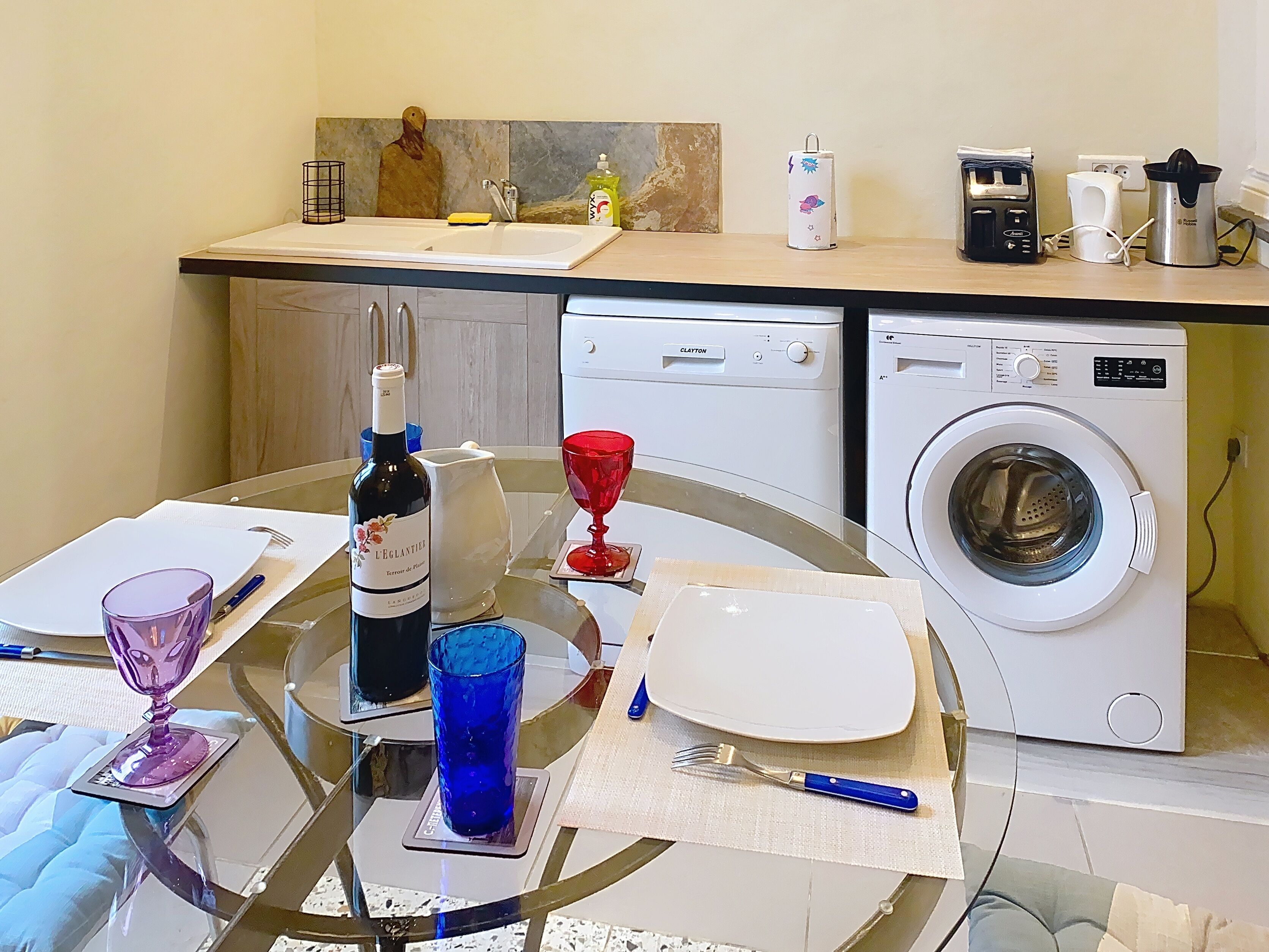 Una cucina con lavatrice, una bottiglia di vino e un tavolo da pranzo apparecchiato con piatti, bicchieri e posate.