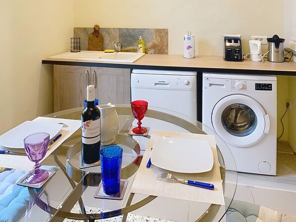 A kitchen with a washing machine, a wine bottle, and a dining table set with plates, glasses, and cutlery. - Résidence Verdi (Bedarieux)