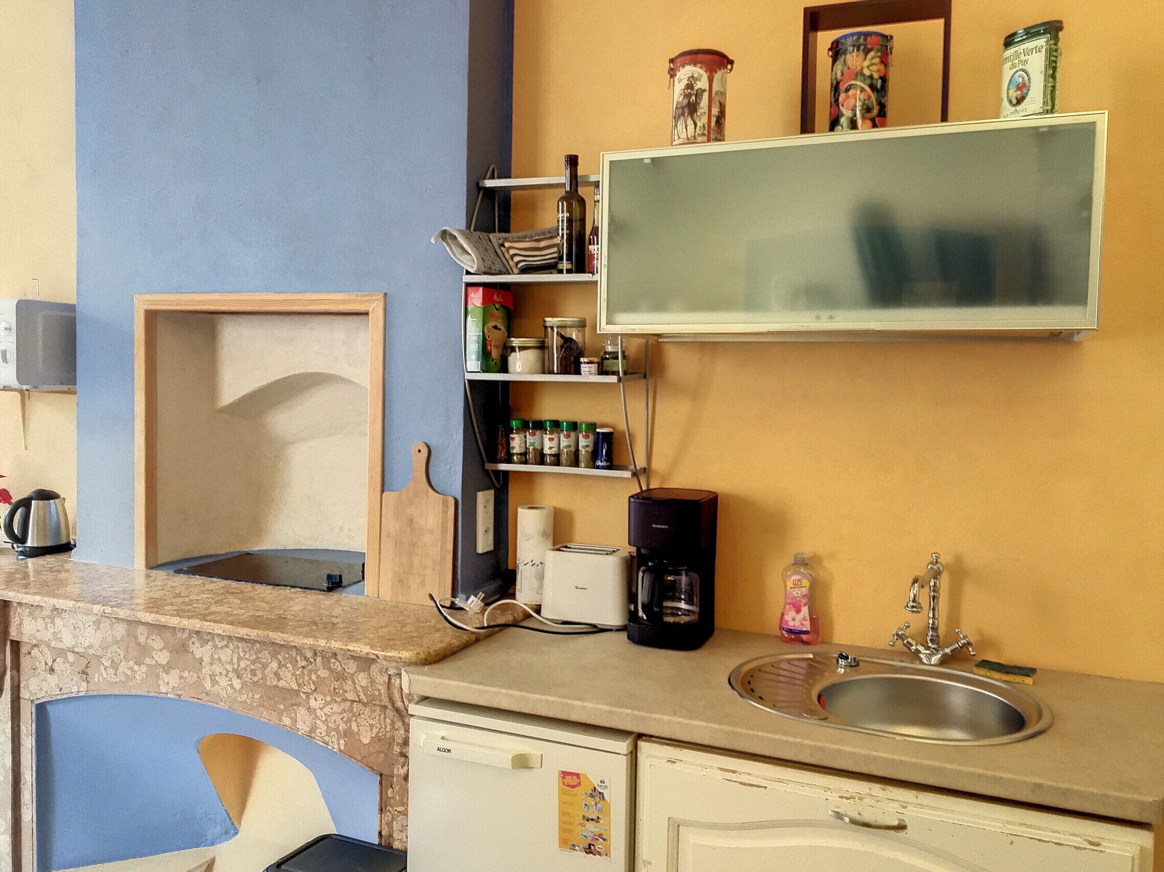 Una cocina compacta con un lavamanos, una cafetera y un microondas. Se aprecia un estante con frascos y un mueble pequeño.