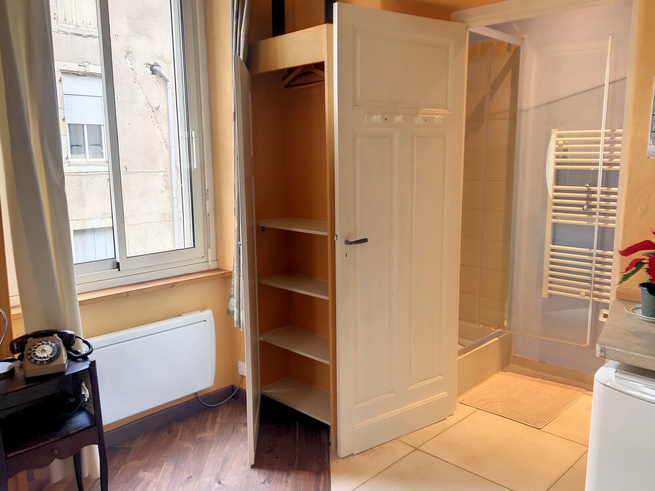 Een kamer met een houten vloer, een radiator, een telefoon, een witte deur en een raam met jaloezieën.