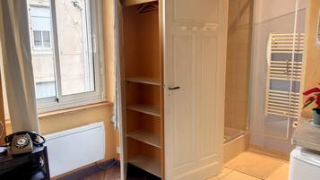 Een kamer met een houten vloer, een radiator, een telefoon, een witte deur en een raam met jaloezieën.