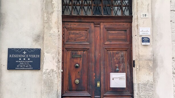 A large wooden double door with a metal grille above it, set in a stone building with a plaque on the wall to the left. - Résidence Verdi (Bedarieux)