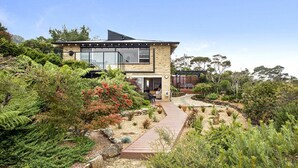 A modern two-story house with a glass conservatory, surrounded by lush vegetation and a wooden pathway. - Donlen Cottage Wentworth Falls 180 Degree Views (WENTWORTH FALLS)