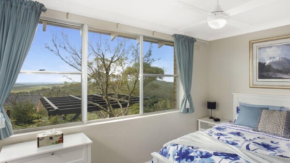 A bedroom with a large window offering a view of a rural landscape, a bed with floral bedding, a bedside table with a lamp, and a framed picture on the wall. - Donlen Cottage Wentworth Falls 180 Degree Views (WENTWORTH FALLS)