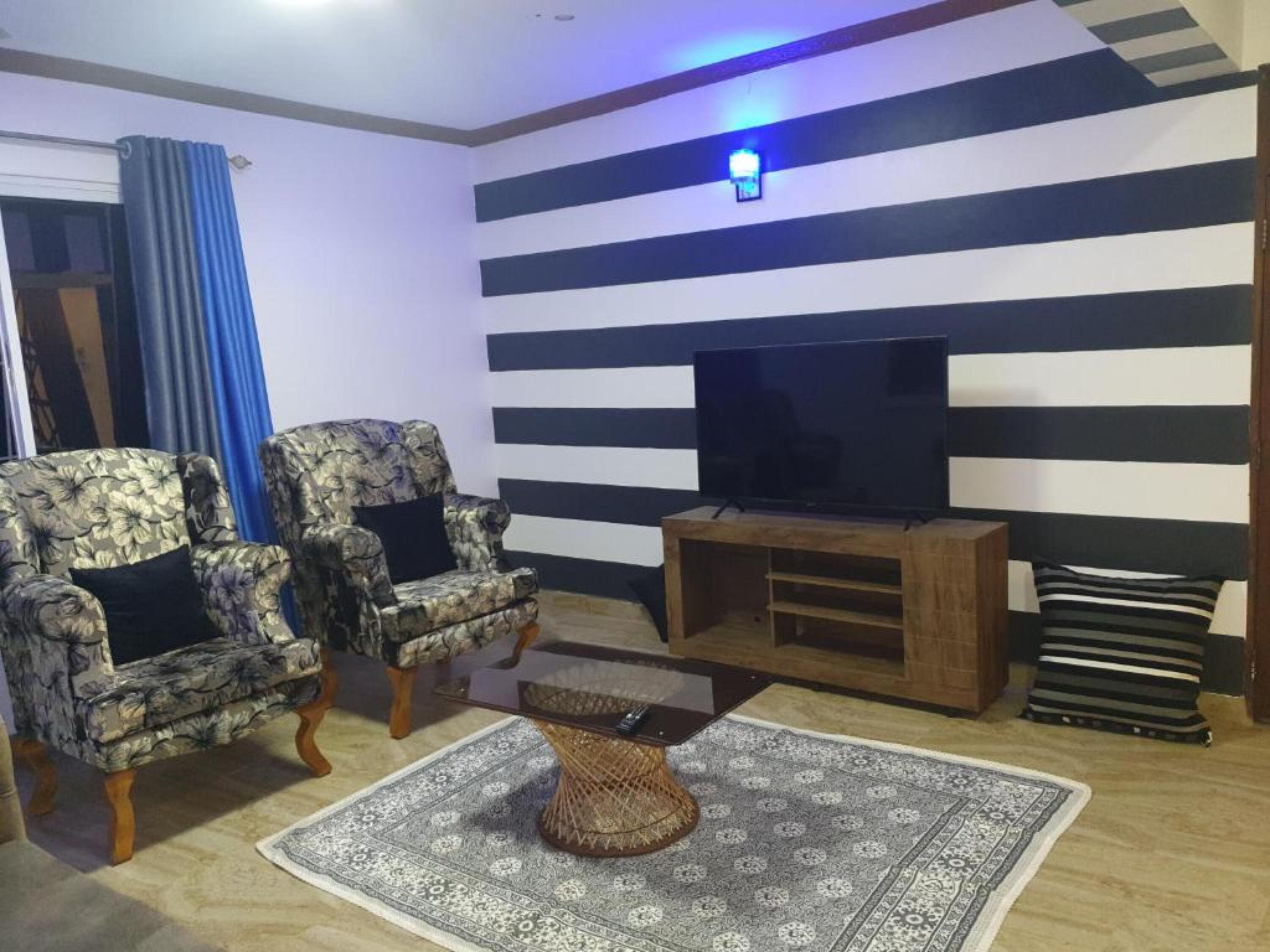 Ein Wohnzimmer mit gestreiften Wänden, einem Flachbildfernseher, zwei gemusterten Sesseln, einem Couchtisch und einem Teppich mit Muster.