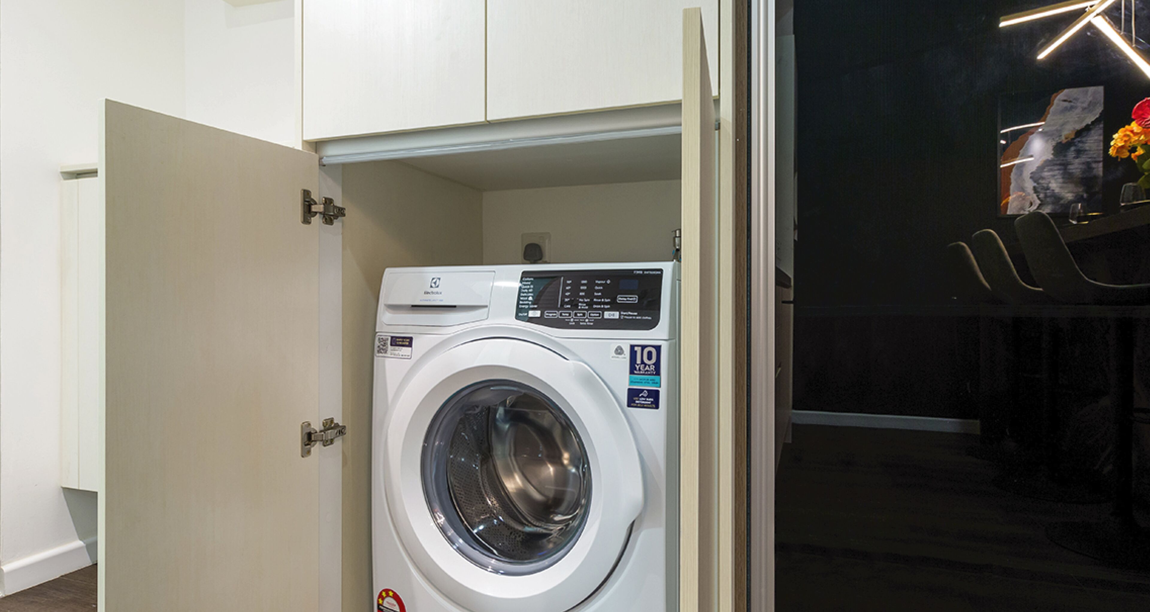 A compact, modern washing machine in a small laundry room with a door leading to a dining area.