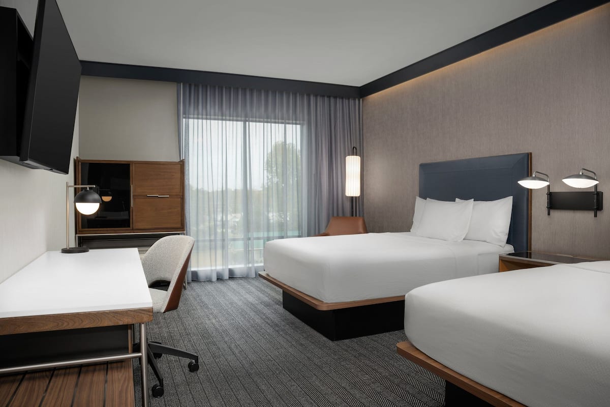 Une chambre d’hôtel avec deux lits, un bureau, une chaise, une télévision et une fenêtre avec des rideaux.