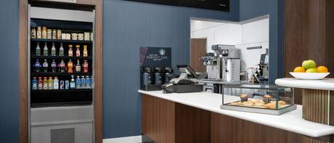 A modern bistro area with a beverage dispenser, coffee machine, and a display of pastries.