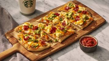 A breakfast pizza with toppings like bacon, avocado, and cheese on a wooden board, a Starbucks cup, and a small bowl of salsa.