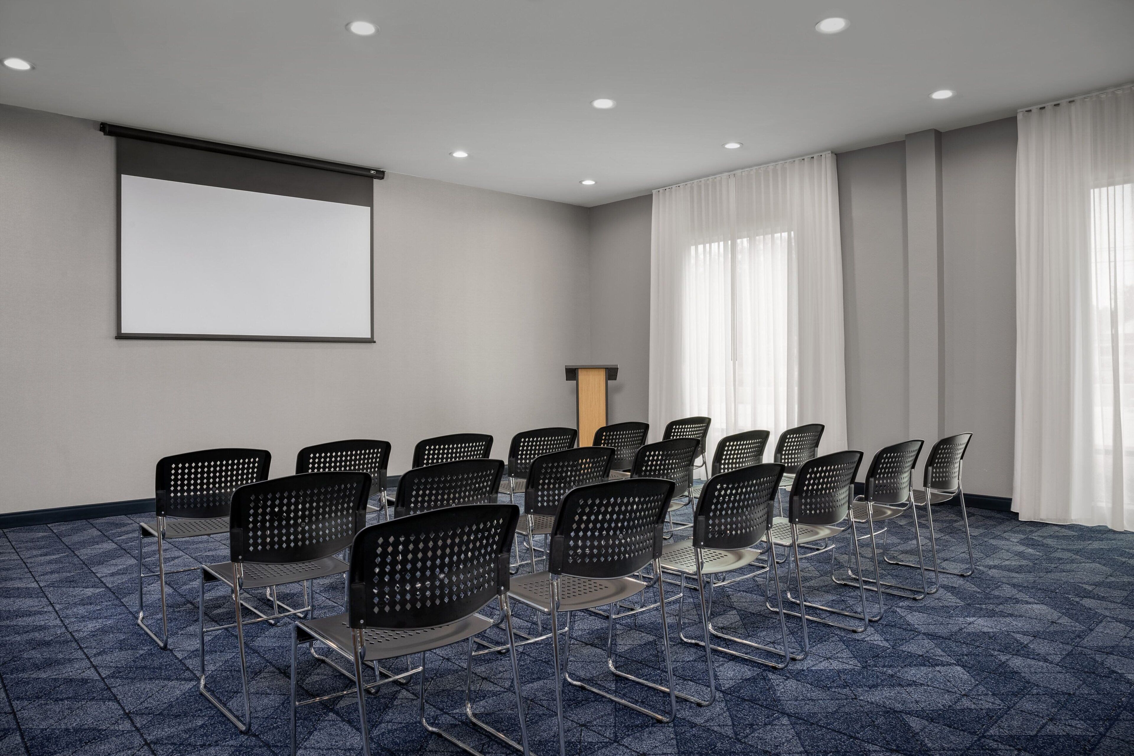 Un salón de conferencias con una pantalla de proyector, filas de sillas, un atril y ventanas amplias con cortinas.