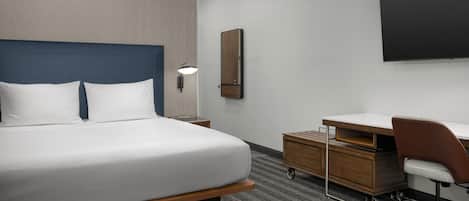 A modern hotel room with a large bed, a desk, a chair, a flat-screen TV, and a small cabinet.
