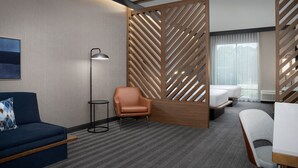 Hotellihuoneessa on sininen sohva, ruskea nojatuoli, pieni pöytä, lattiavalaisin ja sänky, jonka vieressä on puinen väliseinä.