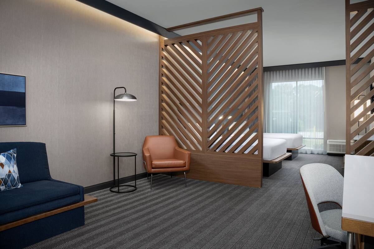 Une chambre d’hôtel avec un canapé bleu, un fauteuil marron, une petite table, un lampadaire et un lit séparé par une cloison en bois.