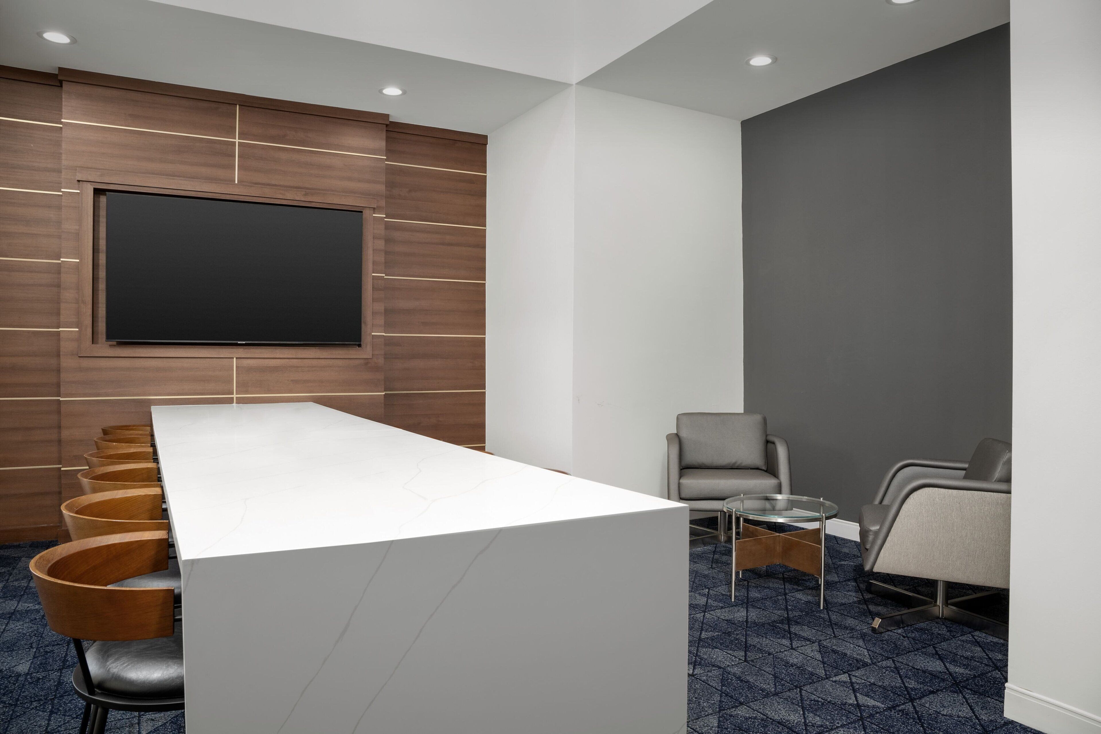 A conference room with a long white table, wooden chairs, a large flat-screen TV, and a seating area with two chairs and a small table.