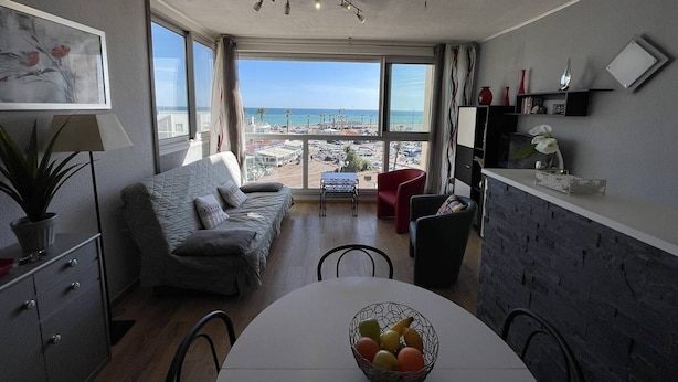 Una sala moderna con un gran ventanal con vista a una playa, una mesa de comedor con fruta y una kitchenette.