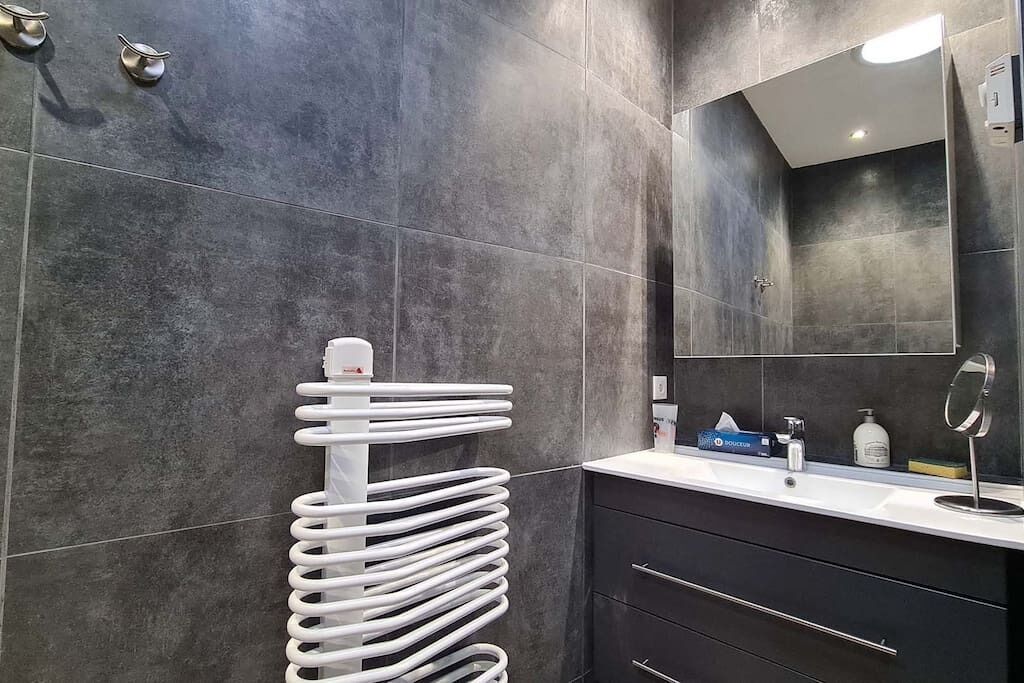 Um banheiro moderno com azulejos escuros, um secador de toalhas branco, um espelho e uma pia com artigos de higiene pessoal.
