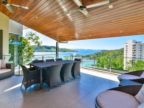 A modern outdoor patio with a dining table and chairs, a sofa, and a view of a body of water and hills.