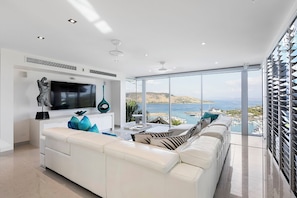 A modern living room with a white sofa, a flat-screen TV, and a view of the sea through large windows.