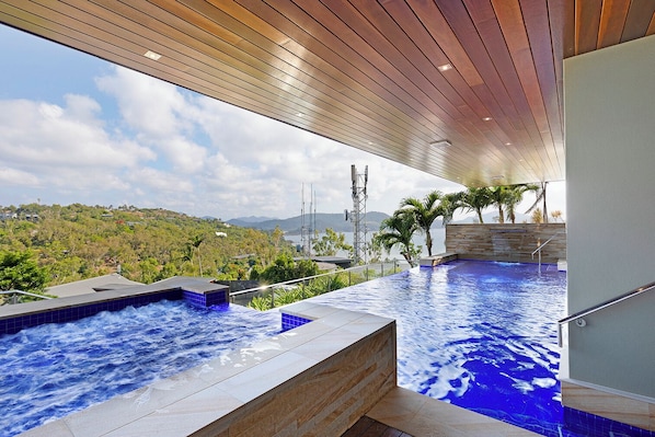 A modern infinity pool with a wooden ceiling, surrounded by lush greenery and a view of distant hills.