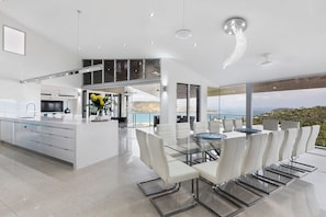 A modern kitchen with a central island, a dining area with a glass table, and a view of the sea.