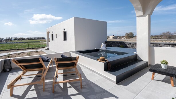A modern outdoor patio with a hot tub, two wooden chairs, and a small table with a plant. - Almira Luxury Suites (Santorini)