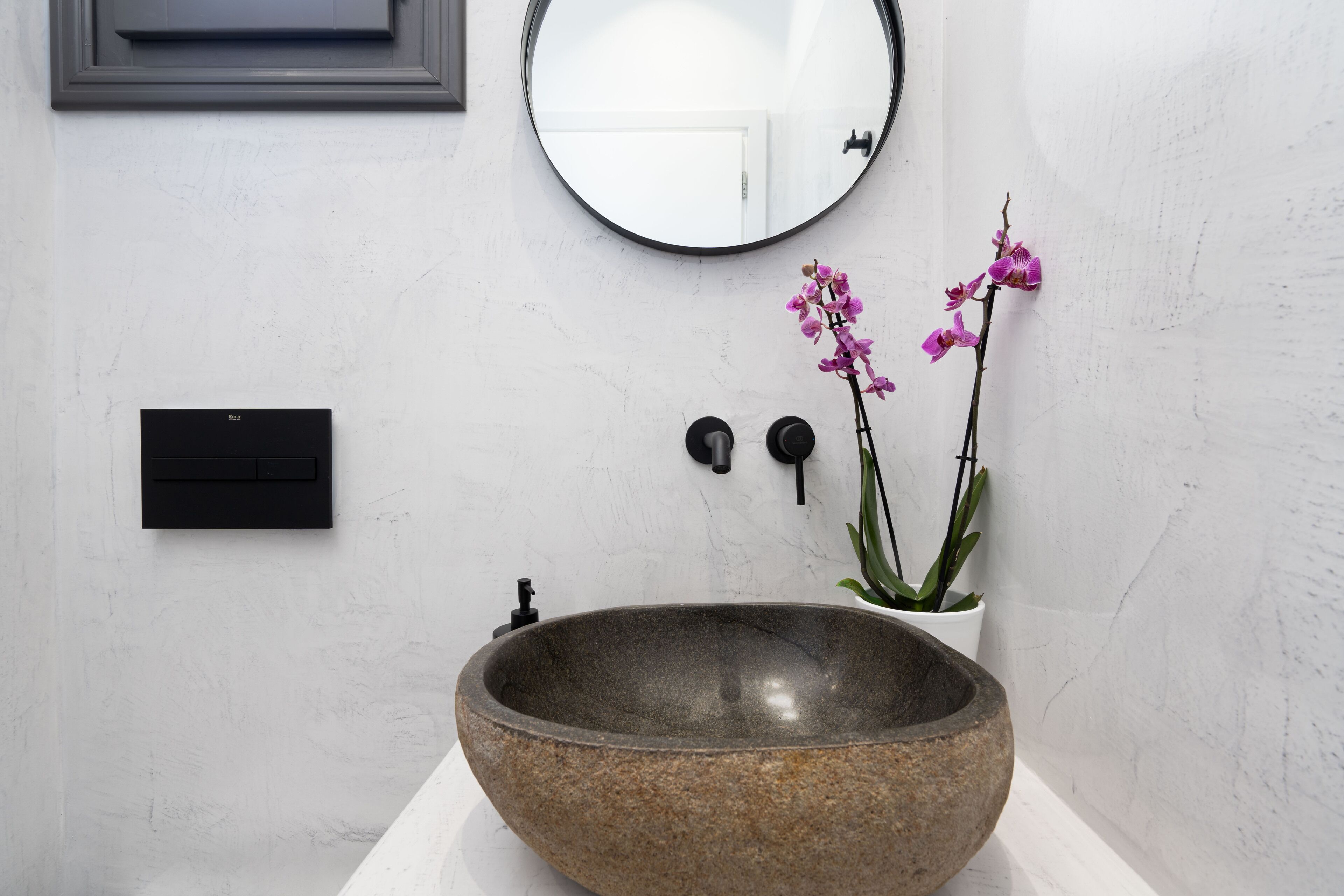 Ein modernes Badezimmer mit einem großen Becken aus Stein, einem runden Spiegel, einer Orchidee im Topf und einem schwarzen Wand-Schalter.