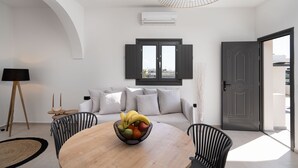 A modern living room with a round wooden table, a bowl of fruit, a sofa with white cushions, a black-framed door, and a ceiling light fixture. - Almira Luxury Suites (Santorini)