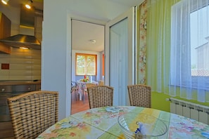 Een eetgedeelte met rieten stoelen, een glazen tafel en uitzicht door een deur op een keuken met een roestvrijstalen afzuigkap.