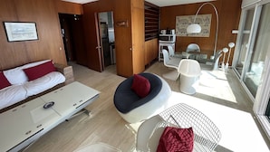 En moderne stue med sofa, spisebord med stole, et køkken og et stort vindue.