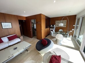 A modern living room with a sofa, a dining table with chairs, a kitchen area, and a large window.