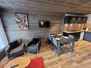 A modern living room with a dining area, grey sofas, a red rug, and a kitchen with dark cabinets and a sink.
