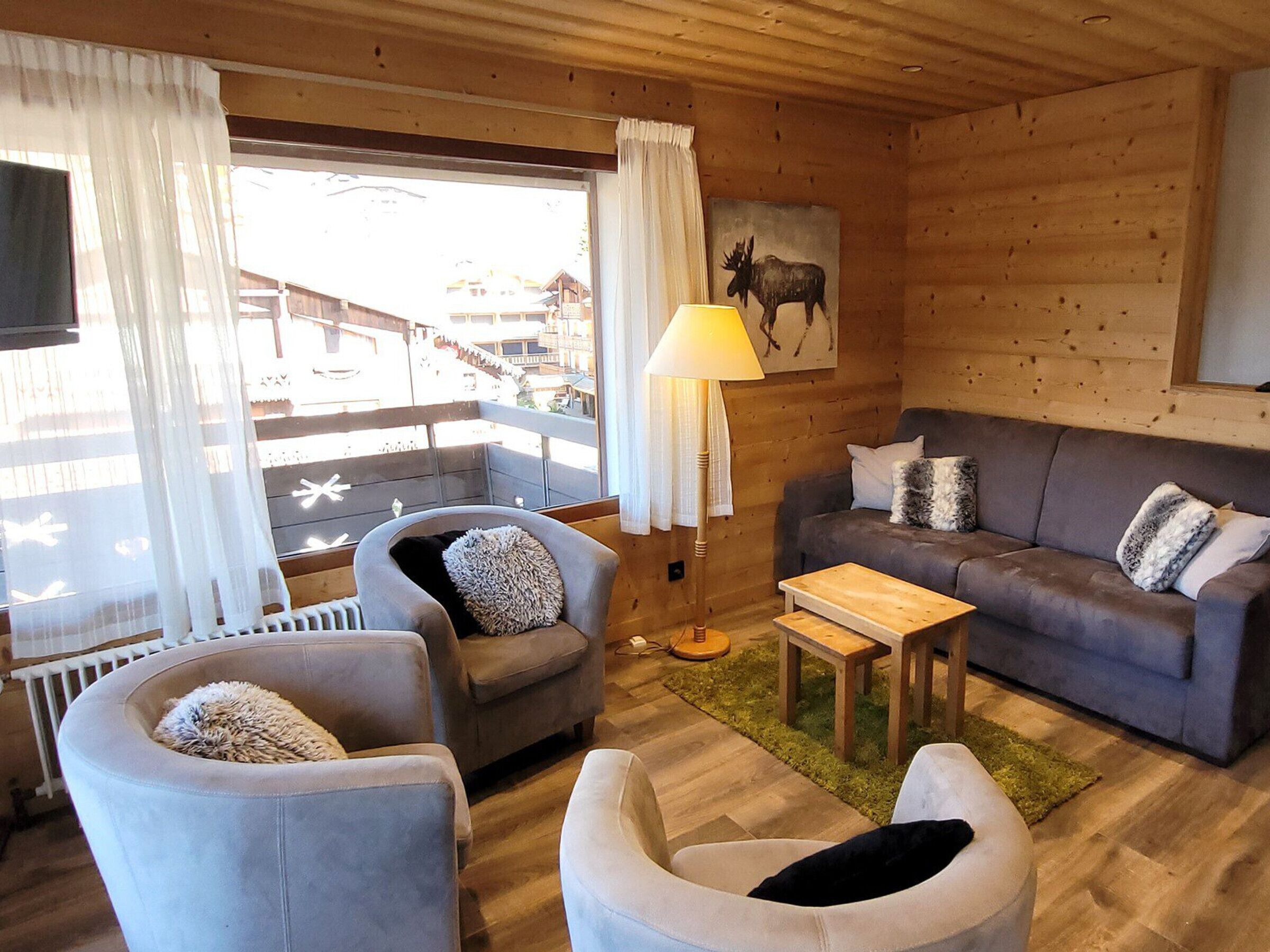 L’intérieur d’un chalet accueillant est aménagé avec un canapé, deux fauteuils, une petite table et une lampe. Un tableau représentant un élan est accroché au mur, et on aperçoit un balcon avec une balustrade.