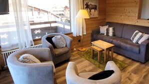A cozy cabin interior with a sofa, two armchairs, a small table, and a lamp. A painting of a moose is on the wall, and there's a view of a balcony with a railing.