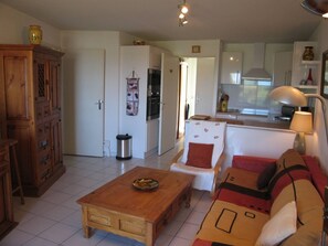 Uma sala de estar com mesa de centro de madeira, sofá com almofadas estampadas e cozinha ao fundo.