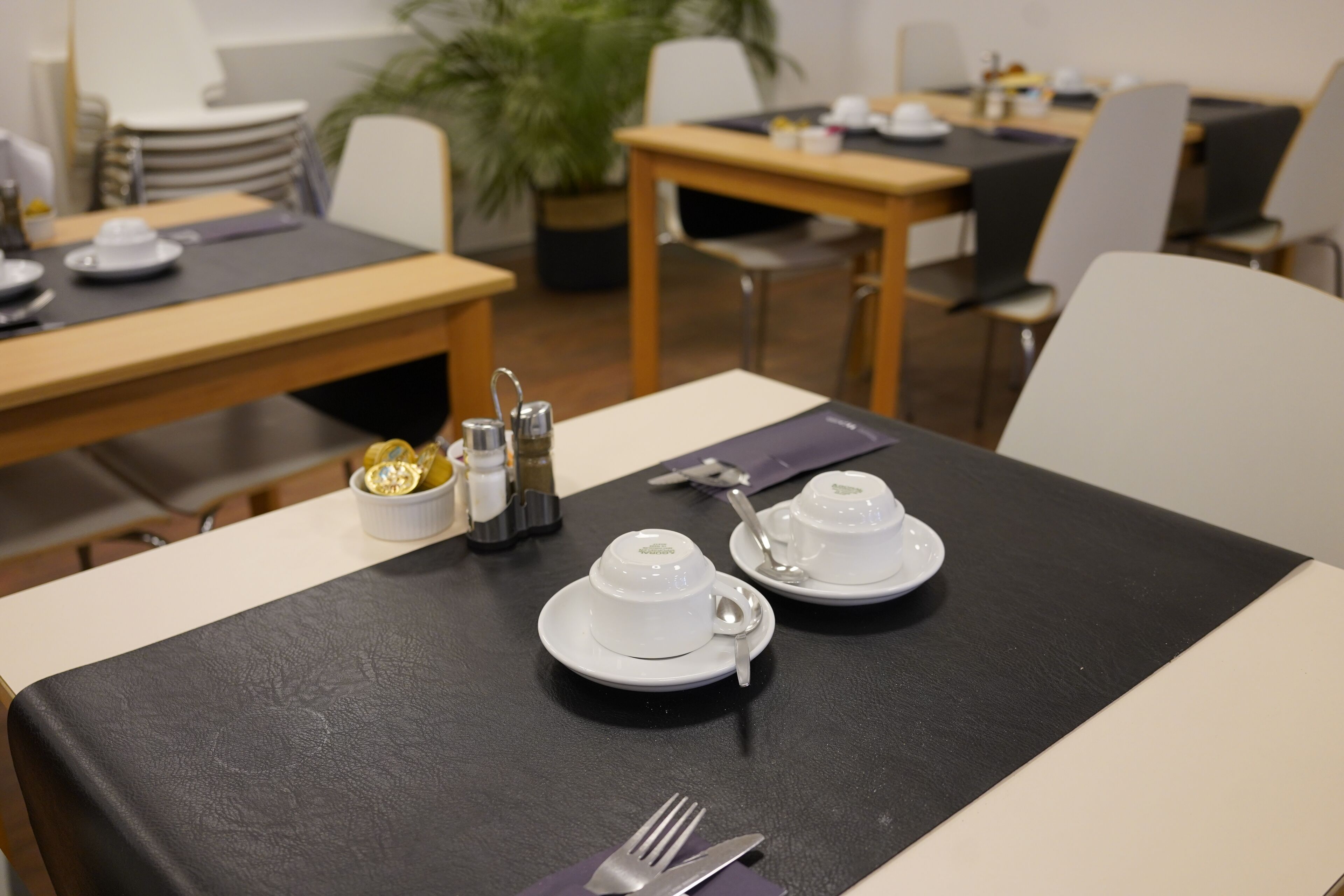 Een eettafel gedekt met witte kopjes, schotels en bestek, met een zwarte tafelonderzetter.