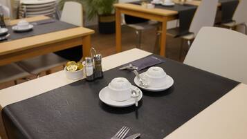 Een eettafel gedekt met witte kopjes, schotels en bestek, met een zwarte tafelonderzetter.