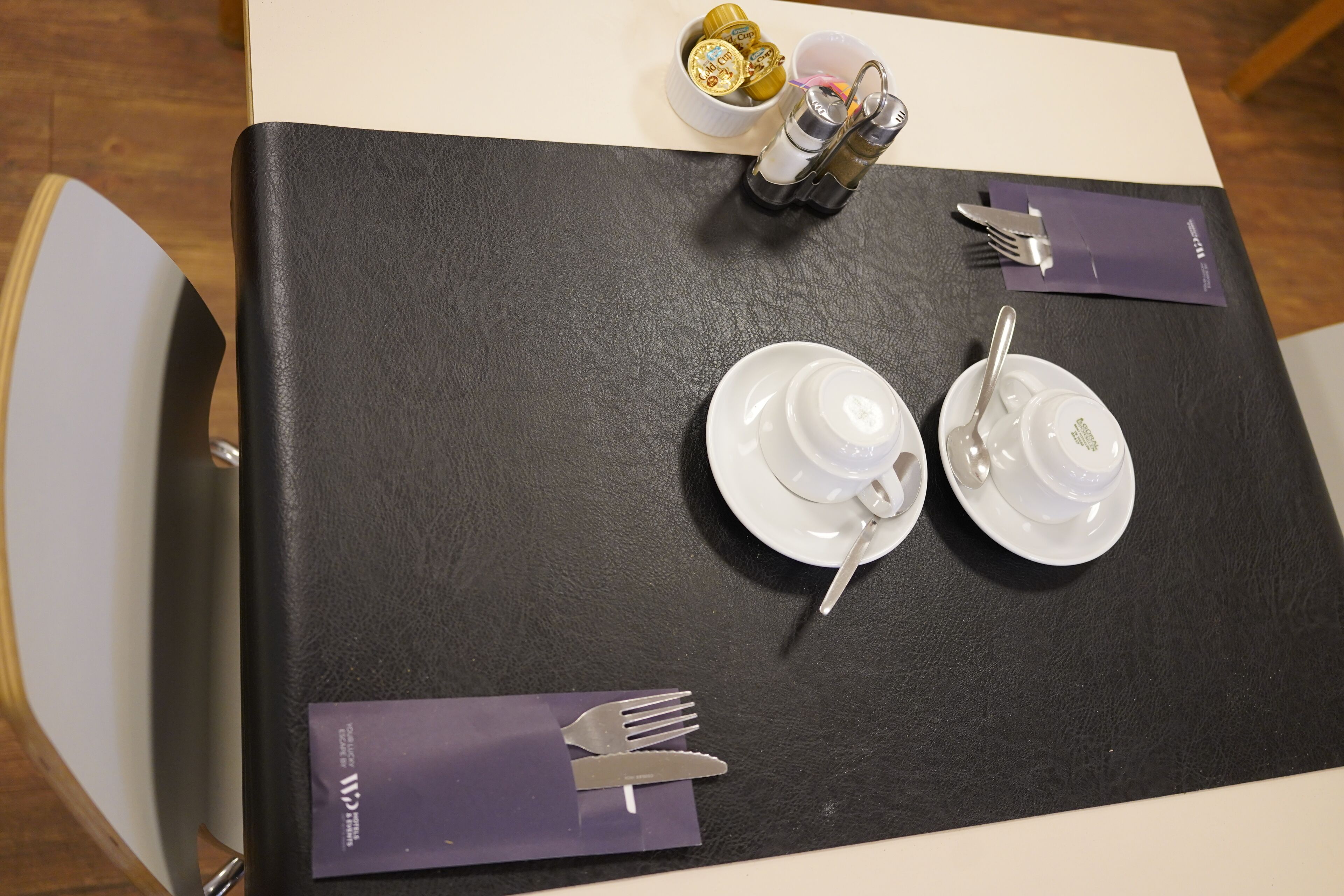 Une table dressée avec de l’assiette blanche, des tasses et des couverts, recouverte d’une nappe noire et agrémentée de serviettes violettes.