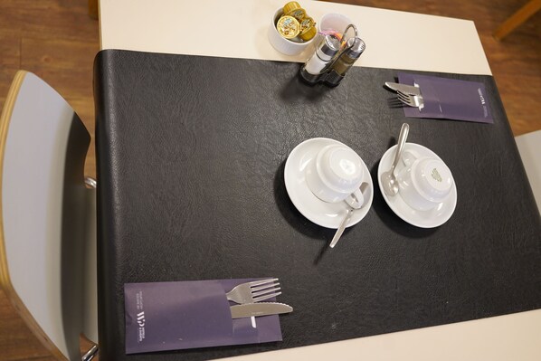 Una mesa dispuesta con platos blancos, tazas y cubiertos, con mantel negro y servilletas moradas.