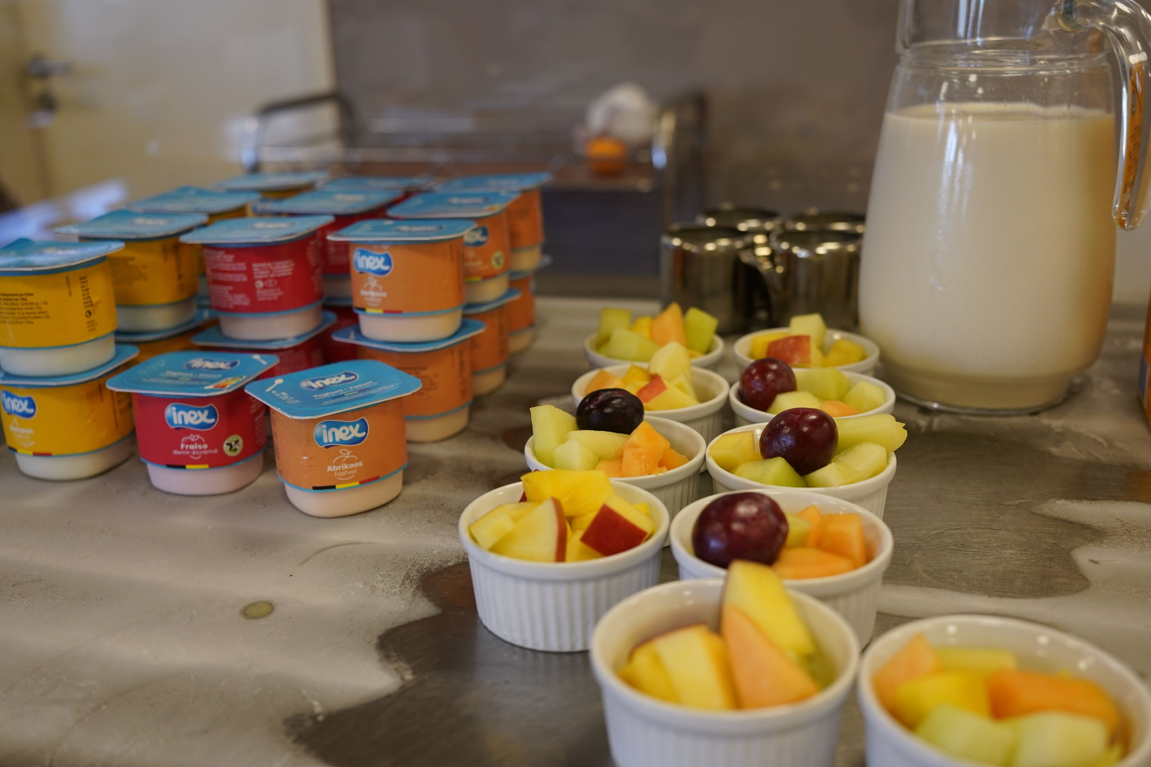 A variety of food items including yogurt containers, fruit bowls, and a pitcher of milk on a stainless steel counter.