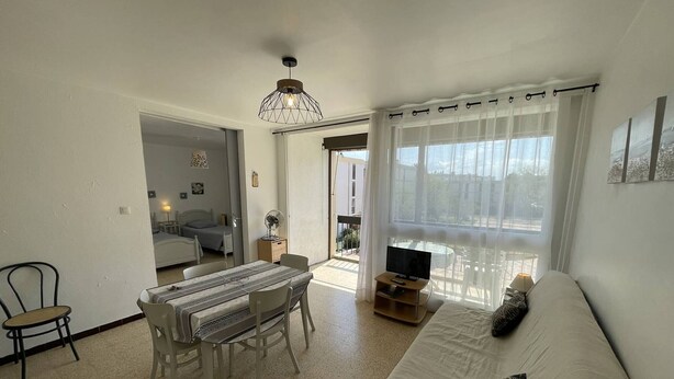 A modern living room with a sofa, dining table, and chairs. There is a TV on a wooden stand and a ceiling light fixture.