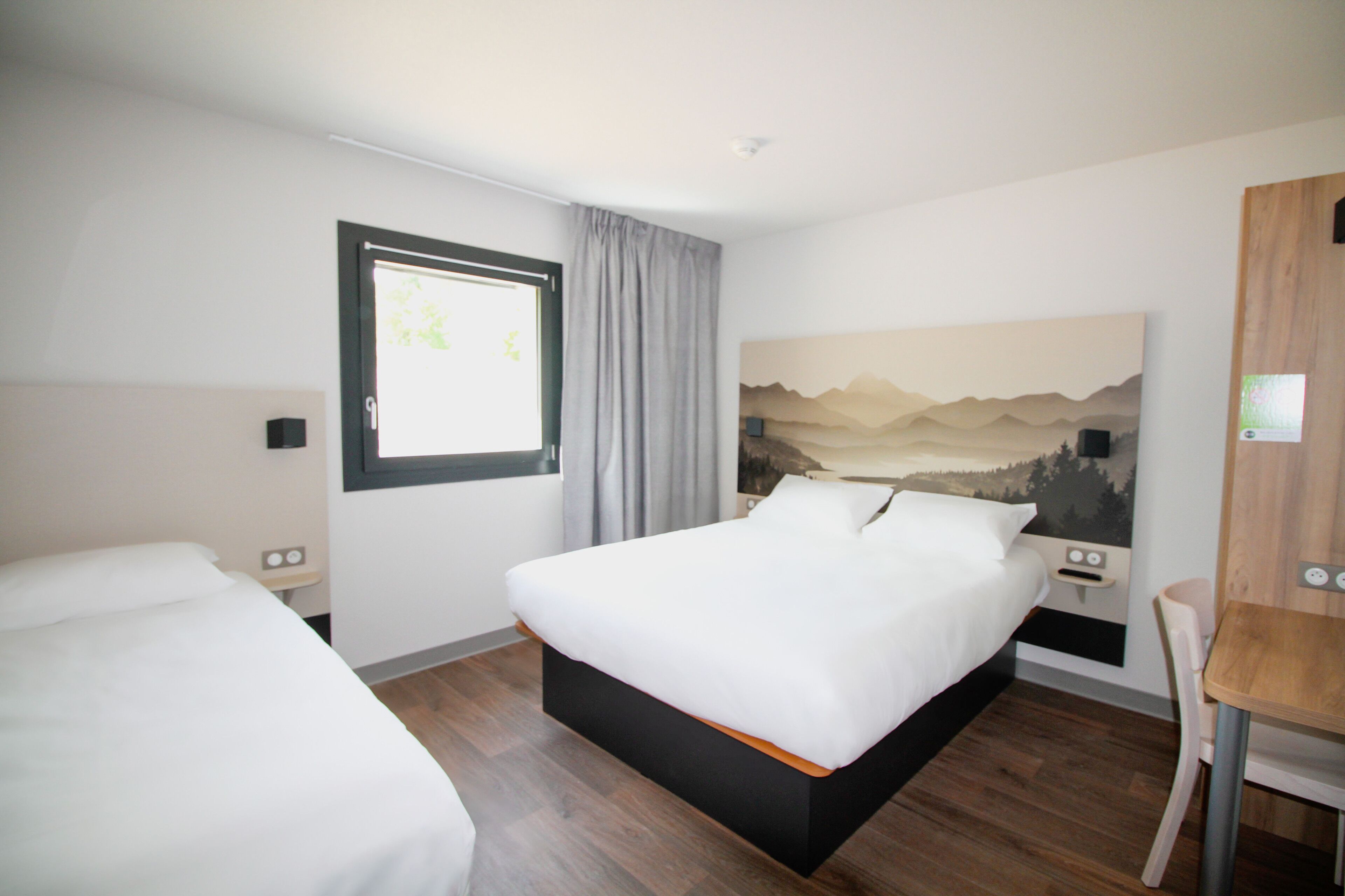 Ein Hotelzimmer mit einem Bett, einem Schreibtisch und einem Stuhl. Ein Fenster mit Vorhängen, ein wandmontierter Fernseher und ein Landschaftsbild sind ebenfalls vorhanden.