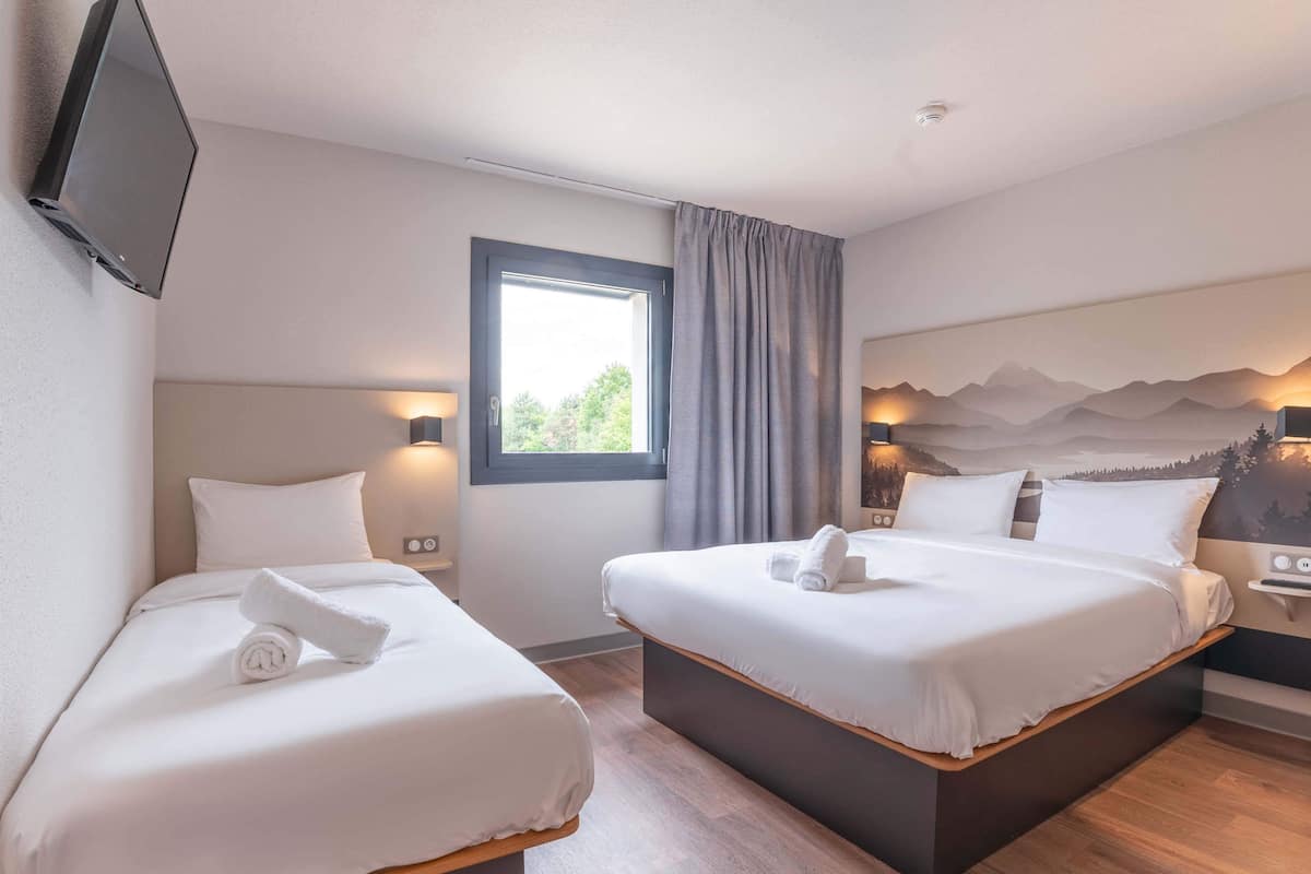 Habitación de hotel con dos camas, un televisor montado en la pared, una ventana con cortinas y una pintura de un paisaje montañoso en la pared.