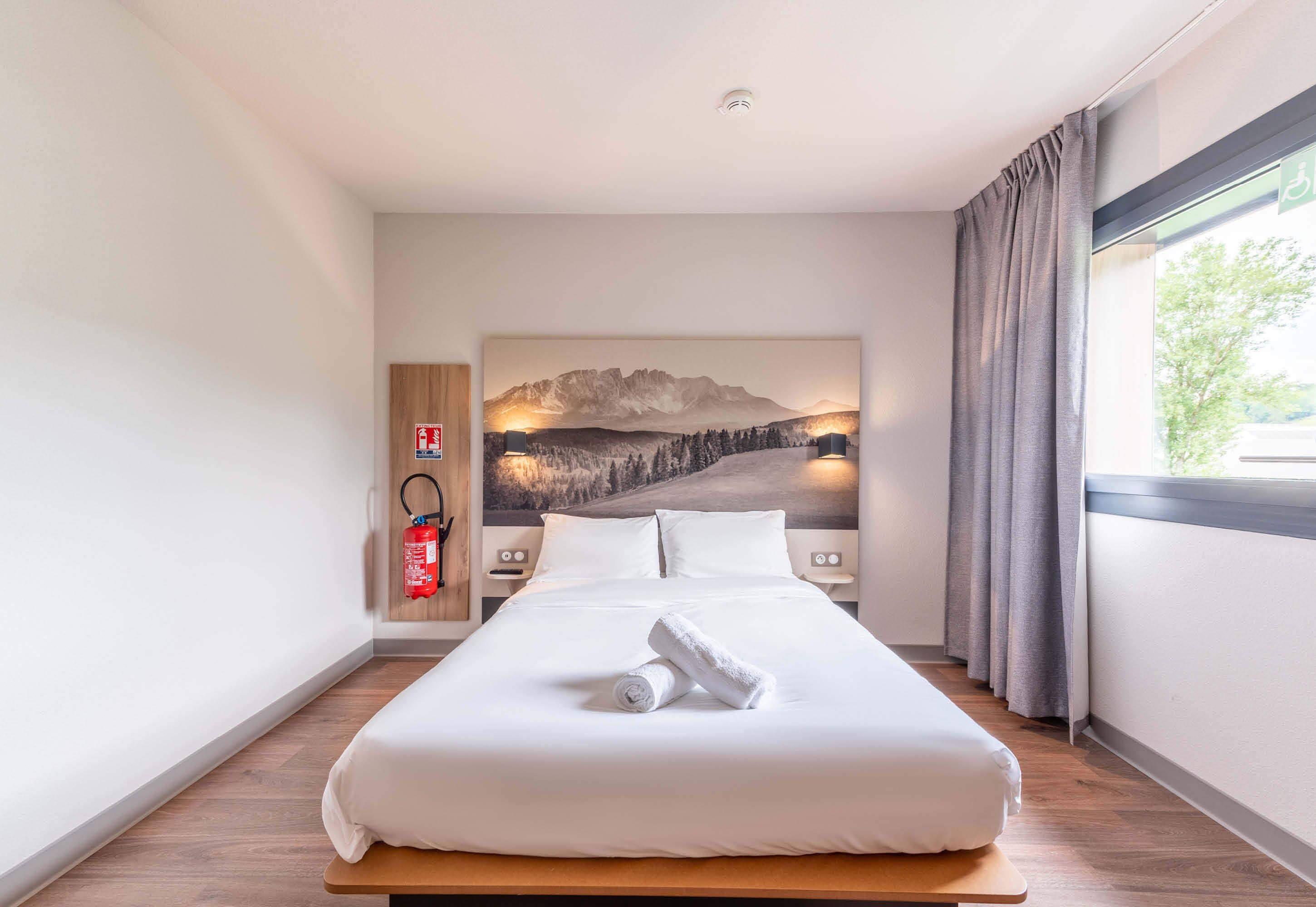 A hotel room with a large bed, a headboard with a mountain landscape painting, a fire extinguisher, and a window with curtains.