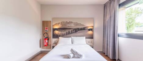 A hotel room with a large bed, a headboard with a mountain landscape painting, a fire extinguisher, and a window with curtains.