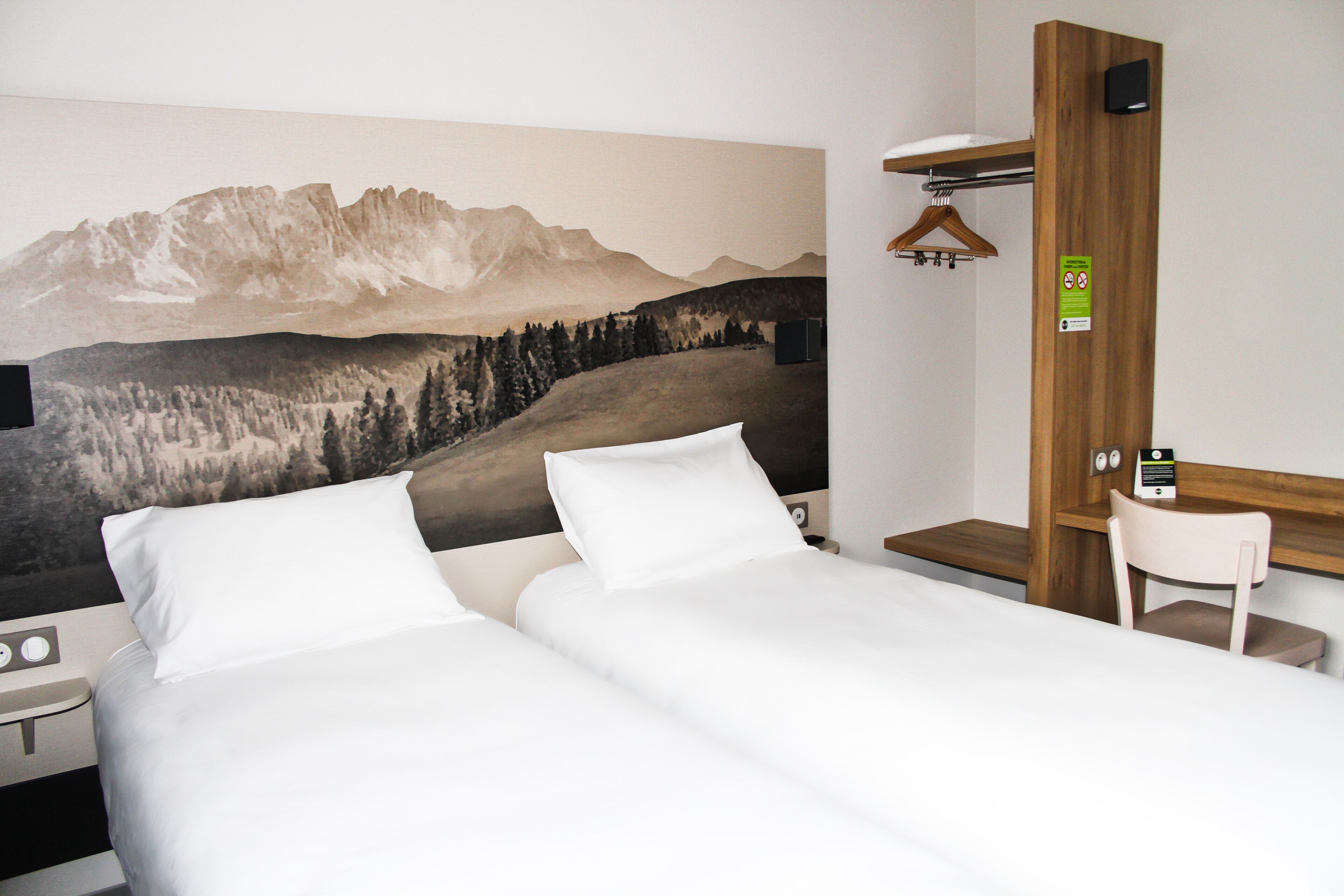 A hotel room with a bed, a desk, and a chair. There is a painting of mountains on the wall.