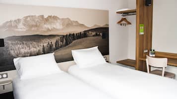 A hotel room with a bed, a desk, and a chair. There is a painting of mountains on the wall.