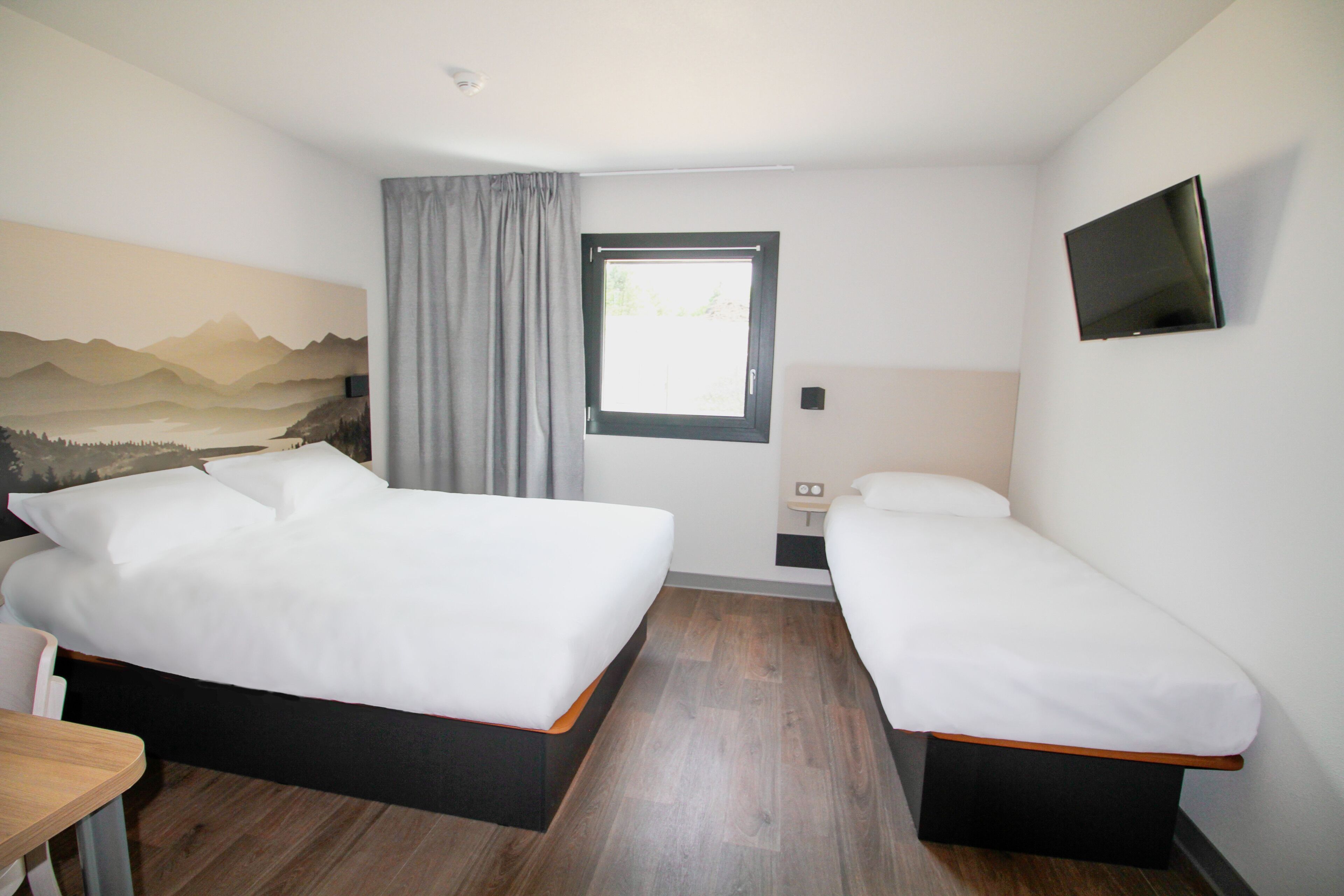 Ein Hotelzimmer mit zwei Betten, einem Schreibtisch, einem Fernseher und einem Fenster mit Vorhängen.