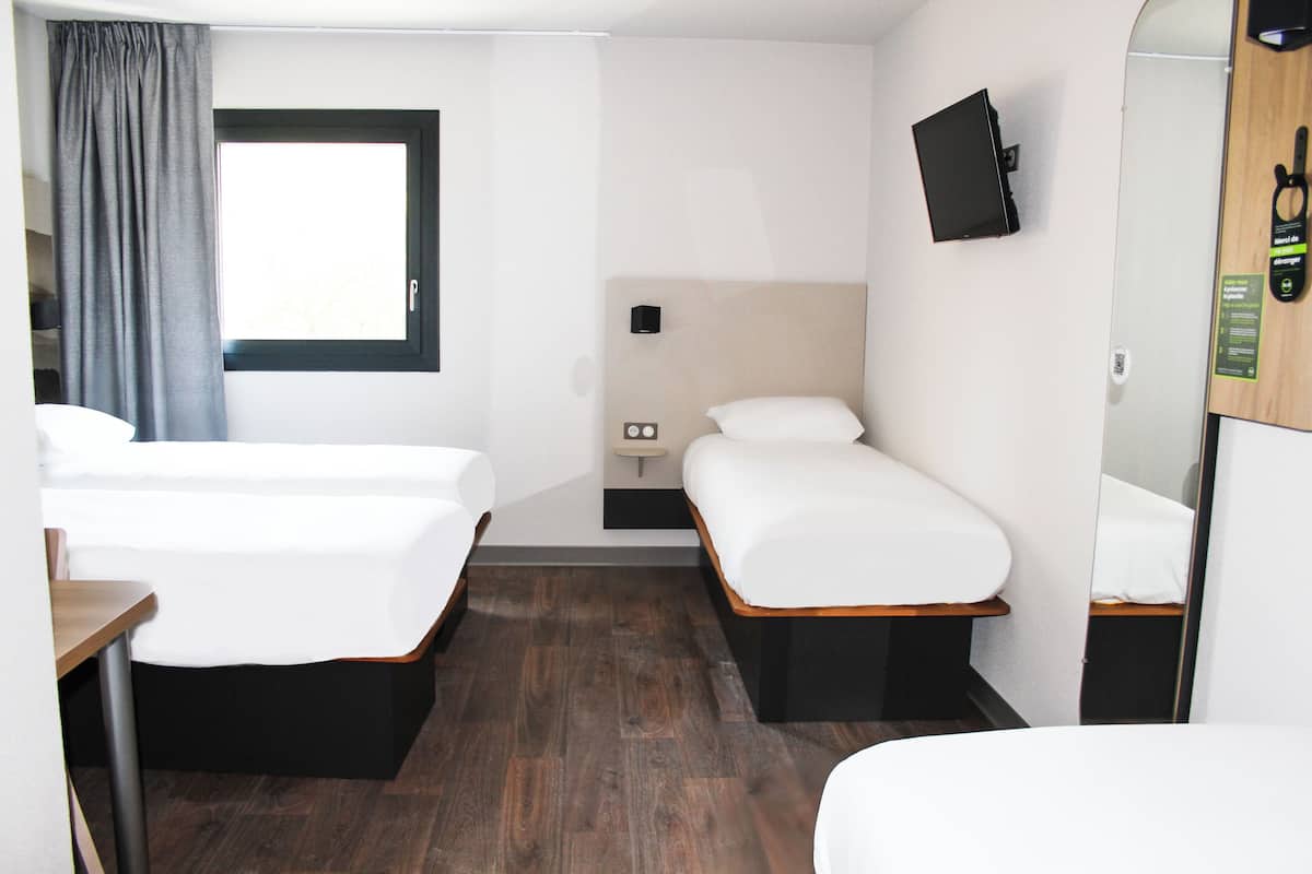 Habitación de hotel con dos camas individuales, una mesita, una ventana con cortinas, un televisor montado en la pared y un espejo.