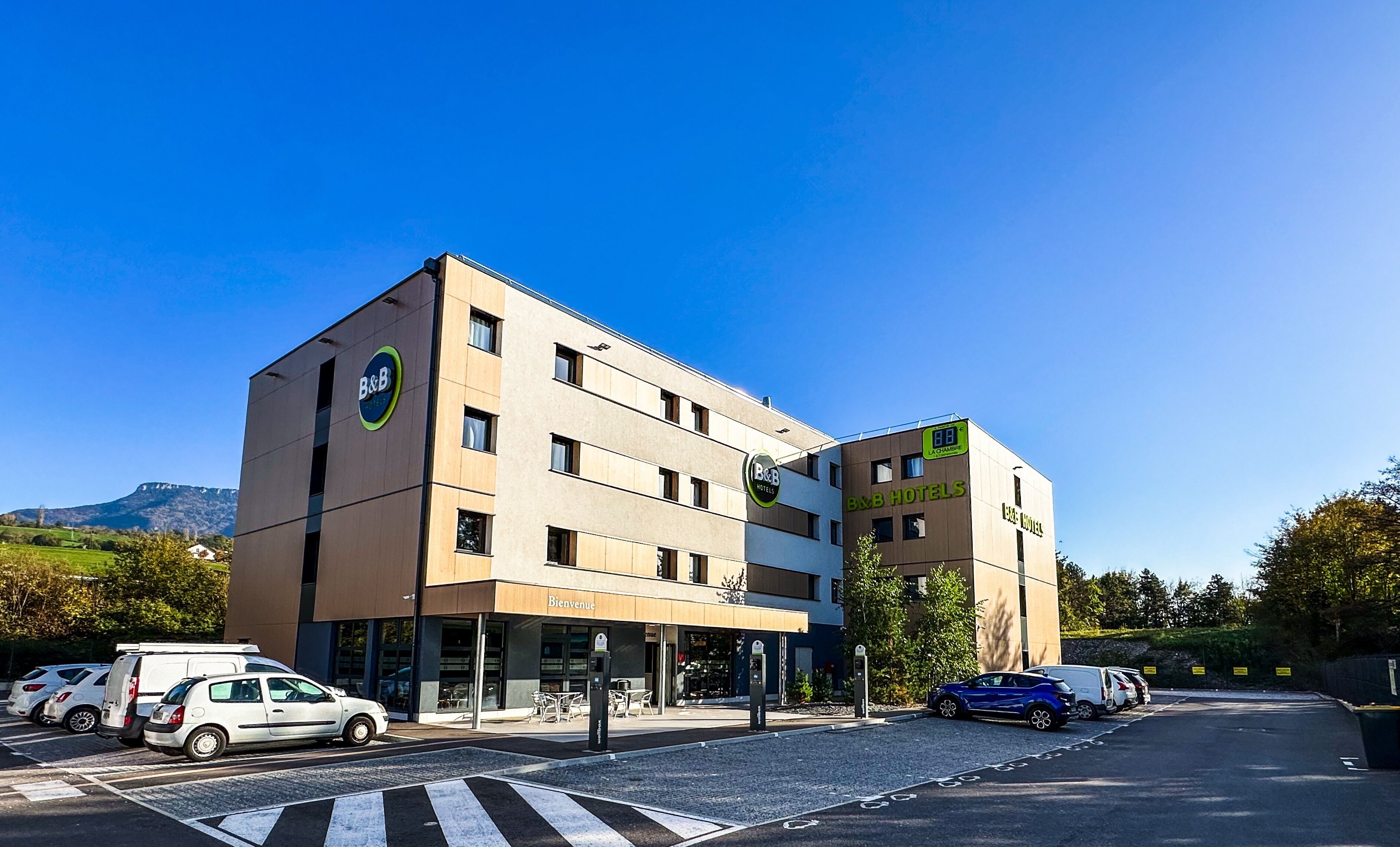 A B&B hotel with a modern design, a visible entrance, and a parking area with cars.